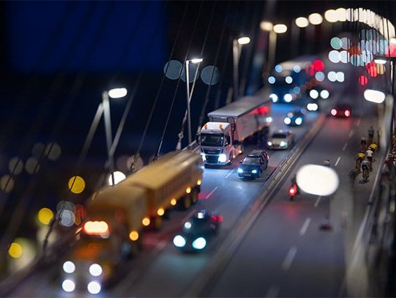 A detailed miniature model of a busy bridge at night, featuring tiny cars, trucks, cyclists, and streetlights