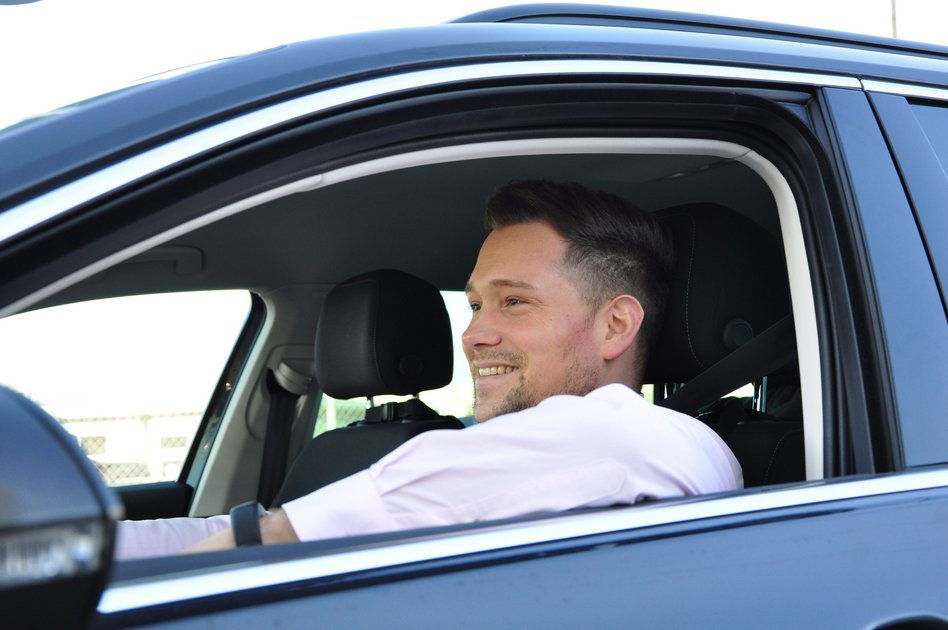 Ein junger Mann mit kurzen dunklen Haaren sitzt in einem schwarzen Auto und lächelt. Er trägt ein helles Hemd und hat beide Hände entspannt am Lenkrad. Das Autofenster ist geöffnet, und im Hintergrund sind ein Zaun und eine offene, helle Umgebung zu sehen. Das Bild vermittelt eine positive und entspannte Atmosphäre im Straßenverkehr.