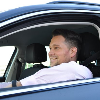Ein junger Mann mit kurzen dunklen Haaren sitzt in einem schwarzen Auto und lächelt. Er trägt ein helles Hemd und hat beide Hände entspannt am Lenkrad. Das Autofenster ist geöffnet, und im Hintergrund sind ein Zaun und eine offene, helle Umgebung zu sehen. Das Bild vermittelt eine positive und entspannte Atmosphäre im Straßenverkehr.