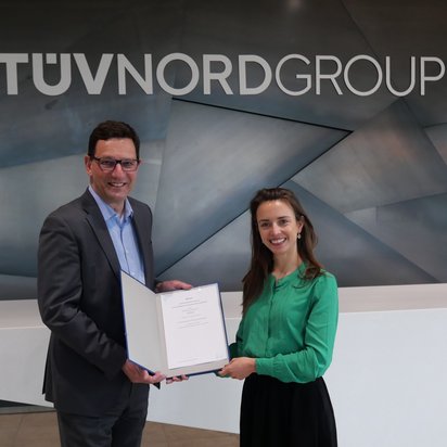 kooperation-tuev-nord-urkundenuebergabe.jpg Ein Mann in einem grauen Anzug mit Brille und eine Frau in einer grünen Bluse stehen vor einer modernen Metallwand mit dem Schriftzug ‚TÜV NORD GROUP‘. Sie halten gemeinsam eine geöffnete Urkunde und lächeln in die Kamera.