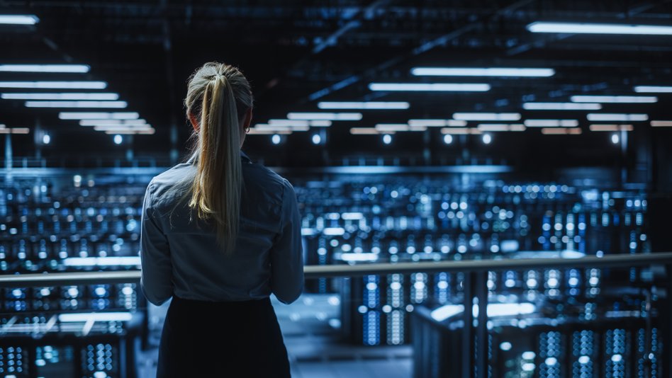 Eine Frau mit langen, blond gebundenen Haaren steht mit dem Rücken zur Kamera und blickt auf ein großes, modern beleuchtetes Rechenzentrum mit zahlreichen Serverracks und leuchtenden LED-Anzeigen.