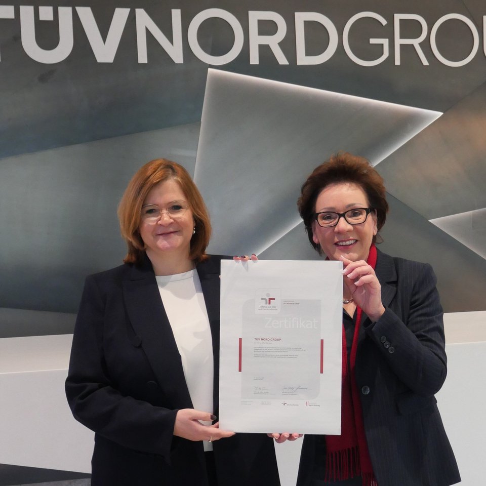 Zwei Frauen stehen vor einer modernen Wand mit dem Logo der TÜV NORD GROUP und halten gemeinsam ein großes Zertifikat. Beide lächeln und tragen Business-Kleidung; die Frau links hat schulterlanges, rötliches Haar und eine dunkle Jacke, die Frau rechts hat kurze, dunkle Haare und trägt eine dunkle Jacke mit rotem Schal. Das Zertifikat ist von der Initiative „berufundfamilie“ und bestätigt das Engagement für eine familienfreundliche Unternehmenskultur. Die Atmosphäre ist professionell und feierlich.