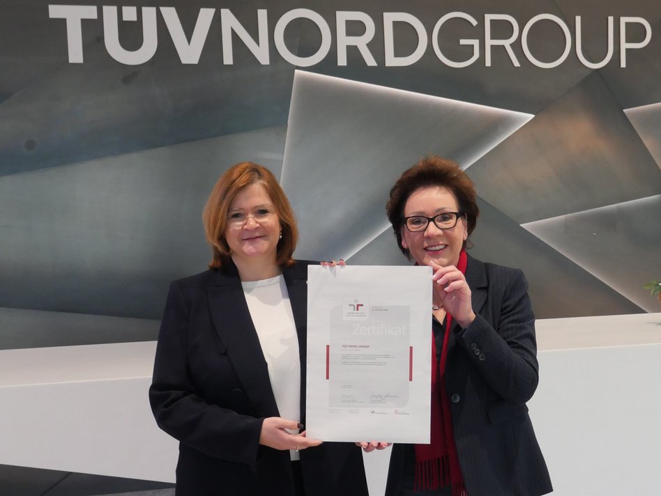 Zwei Frauen stehen vor einer modernen Wand mit dem Logo der TÜV NORD GROUP und halten gemeinsam ein großes Zertifikat. Beide lächeln und tragen Business-Kleidung; die Frau links hat schulterlanges, rötliches Haar und eine dunkle Jacke, die Frau rechts hat kurze, dunkle Haare und trägt eine dunkle Jacke mit rotem Schal. Das Zertifikat ist von der Initiative „berufundfamilie“ und bestätigt das Engagement für eine familienfreundliche Unternehmenskultur. Die Atmosphäre ist professionell und feierlich.