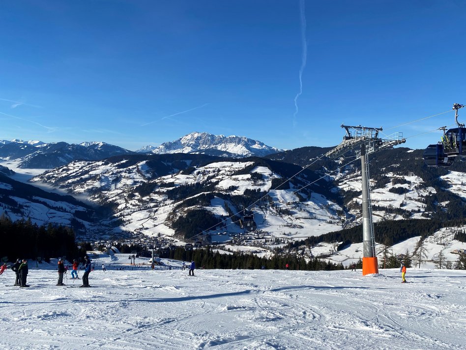 Winterliche Berglandschaft mit schneebedeckten Bergen und einem strahlend blauen Himmel. Im Vordergrund befinden sich mehrere Skifahrer auf einer Piste. Rechts im Bild verläuft eine Gondelbahn mit einer großen Stütze, die eine Seilbahn trägt. Im Hintergrund erstreckt sich ein Tal mit verschneiten Hügeln und Wäldern, während die Sonne die Alpenlandschaft erhellt.