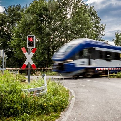Ein Regionalzug mit blau-weißer Lackierung fährt mit hoher Geschwindigkeit über einen Bahnübergang. Die Schranken sind geschlossen, und die roten Warnlichter leuchten. Links und rechts stehen Andreaskreuze zur Kennzeichnung des Bahnübergangs. Die Straße ist von grüner Vegetation umgeben, und im Hintergrund sind Bäume sowie ein bewölkter Himmel zu sehen.