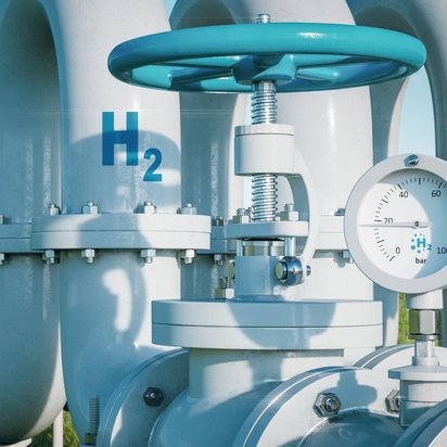 wasserstoff-rohrleitung-ventil-manometer.jpg "Eine große, hellblaue Rohrleitung mit einem Ventil und einem Manometer im Vordergrund. Auf der Rohrleitung ist deutlich die chemische Formel 'H2' für Wasserstoff zu sehen. Im unscharfen Hintergrund befinden sich moderne weiße Gebäude, eine grüne Wiese, ein Windrad und ein klarer blauer Himmel. Das Manometer zeigt '2 bar' an.