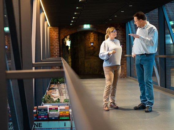 A man and woman converse on a modern walkway overlooking a detailed miniature shipping container model.