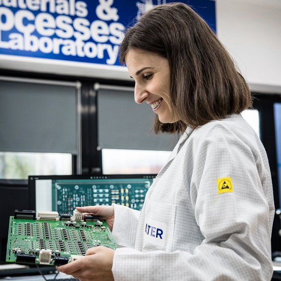 Eine Ingenieurin in einem weißen ESD-Schutzkittel untersucht eine Leiterplatte.